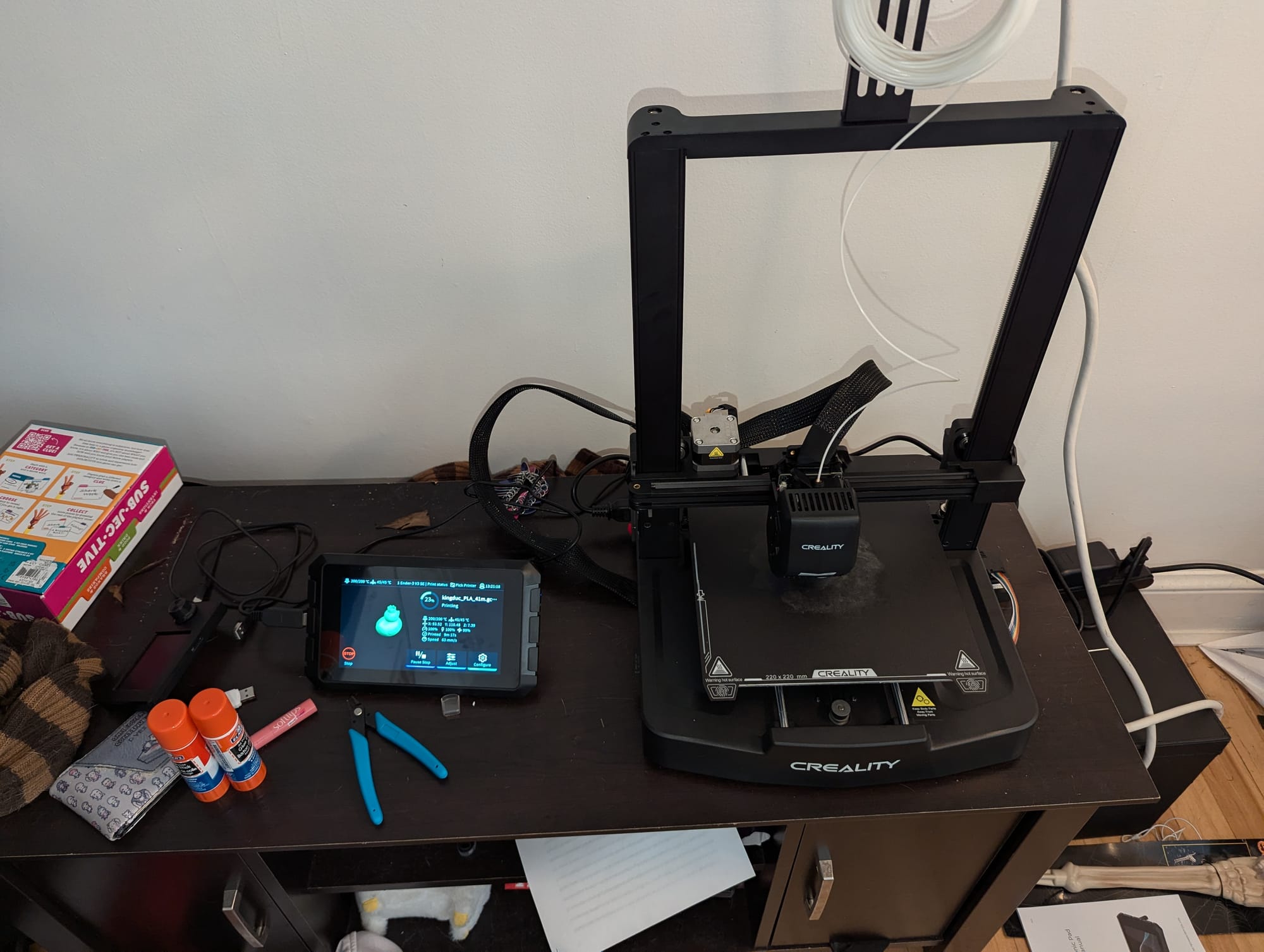 Photo of the Creality Ender 3 V3 SE, with the Creality Sonic Pad to the left of it on top of a repurposed TV stand. The screen shows the current print: a small duck with a crown.